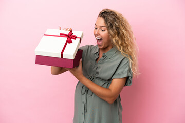 Girl with curly hair isolated on pink background pregnant and holding a gift with surprised expression