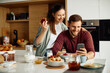 Happy couple use mobile phone while having breakfast together in the morning.