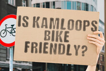 The Question " Is Kamloops Bike Friendly? " On A Banner In Men's Hand With Blurred Background. Transportation. Zero Waste. Bicycle Lane. Streets. City. Safety. Insecure. Road Signs. Dangerous