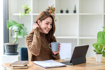 Wall Mural - Beautiful young Asian woman smiling holding a coffee mug and laptop, tablet working at the office..