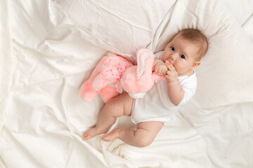 cheerful newborn baby in a white bodysuit lies on a white bed with a plush pink toy rabbit. products for children, toy. concept of a happy childhood and motherhood. child care. space for text.