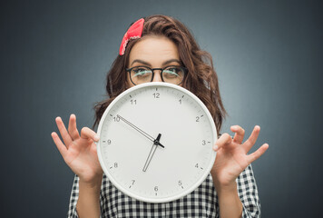 Astonished young woman with surprised face holding clock over dark gray - black background. Pretty girl showing wall clock.