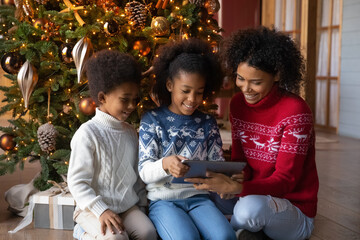 Wall Mural - Happy young African American mother and two little kids sit near Christmas tree play on modern tablet. Smiling biracial mom and small children have fun using pad gadget on winter holidays at home.