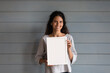 © fizkes - Happy pretty Hispanic 30s woman with curly hair holding blank empty white picture frame with copy space for text, standing at grey wall background, looking at camera. Head shot portrait