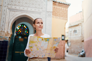 Wall Mural - Travel and active lifestyle concept. Young traveller woman walking in ancient moroccan town holding tourist map.