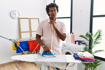 Poster - African man with curly hair ironing clothes at home thinking concentrated about doubt with finger on chin and looking up wondering