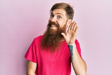 Canvas Print - Redhead man with long beard wearing casual pink t shirt smiling with hand over ear listening and hearing to rumor or gossip. deafness concept.