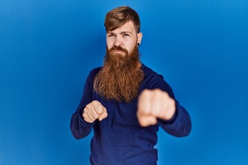 Poster - Redhead man with long beard wearing casual blue sweater over blue background punching fist to fight, aggressive and angry attack, threat and violence