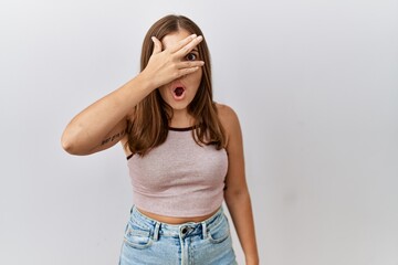 Canvas Print - Young brunette woman standing over isolated background peeking in shock covering face and eyes with hand, looking through fingers with embarrassed expression.