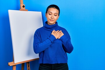 Canvas Print - Beautiful hispanic woman with short hair standing by painter easel stand smiling with hands on chest with closed eyes and grateful gesture on face. health concept.