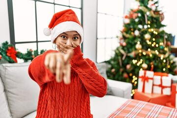 Sticker - Young hispanic woman with short hair wearing christmas hat sitting on the sofa laughing at you, pointing finger to the camera with hand over mouth, shame expression