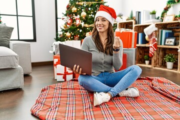 Young latin woman using laptop sitting by christmas tree doing money gesture with hands, asking for salary payment, millionaire business