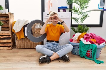 Poster - Young blonde woman doing laundry sitting by washing machine doing heart shape with hand and fingers smiling looking through sign