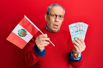 Canvas Print - Handsome senior man with grey hair holding peru flag and peruvian sol banknotes clueless and confused expression. doubt concept.