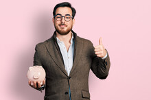 Young Hispanic Man Holding Piggy Bank Smiling Happy And Positive, Thumb Up Doing Excellent And Approval Sign