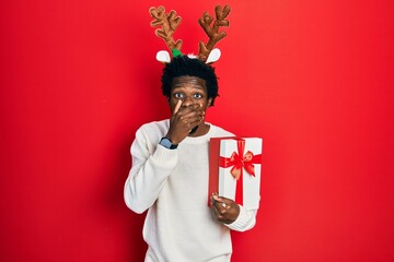 Canvas Print - Young african american man wearing deer christmas hat holding gift shocked covering mouth with hands for mistake. secret concept.