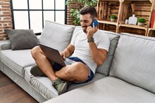 Young hispanic man using laptop and talking on the smartphone at home