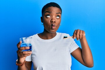 Poster - Young african american woman holding glass of mouthwash and toothbrush for fresh breath making fish face with mouth and squinting eyes, crazy and comical.