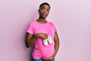 Poster - Young african american woman expecting a baby holding shoes relaxed with serious expression on face. simple and natural looking at the camera.