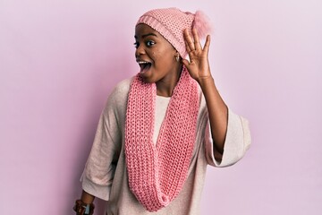 Poster - Young african american woman wearing wool hat and winter scarf smiling with hand over ear listening and hearing to rumor or gossip. deafness concept.