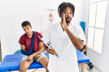 Canvas Print - Young hispanic man working at pain recovery clinic with a man with broken arm hand on mouth telling secret rumor, whispering malicious talk conversation