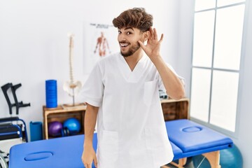 Wall Mural - Young arab man working at pain recovery clinic smiling with hand over ear listening an hearing to rumor or gossip. deafness concept.