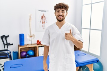 Wall Mural - Young arab man working at pain recovery clinic doing happy thumbs up gesture with hand. approving expression looking at the camera showing success.