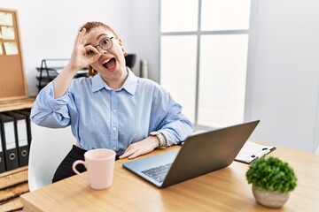 Sticker - Young redhead woman working at the office using computer laptop doing ok gesture with hand smiling, eye looking through fingers with happy face.