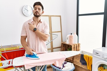 Poster - Young man with beard ironing clothes at home pointing with hand finger to the side showing advertisement, serious and calm face