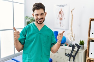 Poster - Young man with beard working at pain recovery clinic success sign doing positive gesture with hand, thumbs up smiling and happy. cheerful expression and winner gesture.