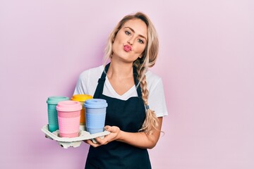 Poster - Beautiful young blonde woman wearing waitress apron holding take away cup of coffees looking at the camera blowing a kiss being lovely and sexy. love expression.