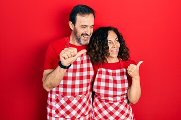 Wall Mural - Middle age couple of hispanic woman and man wearing cook apron smiling with happy face looking and pointing to the side with thumb up.