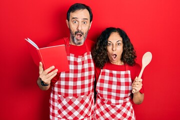 Wall Mural - Middle age couple of hispanic woman and man wearing professional apron reading cooking recipe book afraid and shocked with surprise and amazed expression, fear and excited face.