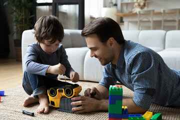 Concentrated small preschool kid boy using screwdriver, learning fixing toy car with caring smiling young father, improving technical skills, playing together on weekend in modern living room.