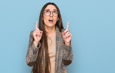 Young hispanic girl wearing business clothes and glasses amazed and surprised looking up and pointing with fingers and raised arms.