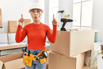 Canvas Print - Middle age grey-haired woman wearing hardhat standing at new home amazed and surprised looking up and pointing with fingers and raised arms.