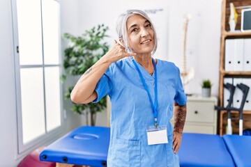 Poster - Middle age grey-haired woman wearing physiotherapist uniform at medical clinic smiling doing phone gesture with hand and fingers like talking on the telephone. communicating concepts.