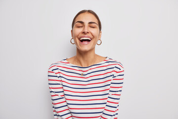 Wall Mural - Indoor shot of happy woman keeps eyes clsed smiles broadly shows teeth laughs at something positive dressed in casual striped jumper isolated over white backgroud. People and emotions concept