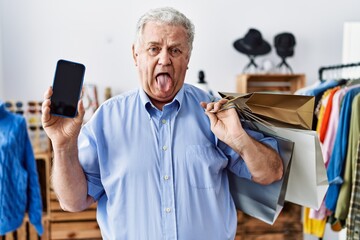 Sticker - Senior man with grey hair holding shopping bags and showing smartphone screen sticking tongue out happy with funny expression.