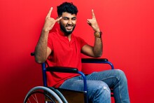 Arab Man With Beard Sitting On Wheelchair Shouting With Crazy Expression Doing Rock Symbol With Hands Up. Music Star. Heavy Music Concept.