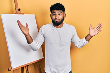 Canvas Print - Arab man with beard standing by painter easel stand clueless and confused with open arms, no idea and doubtful face.