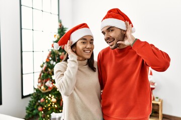 Poster - Young hispanic couple standing by christmas tree smiling with hand over ear listening an hearing to rumor or gossip. deafness concept.