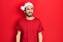 Young Hispanic Man Wearing Christmas Hat Looking Away To Side With Smile On Face, Natural Expression. Laughing Confident.