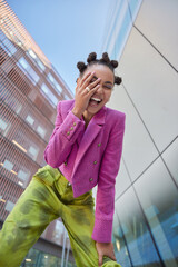View from below of overjoyed female model keeps hand on face feels very happy wears stylish pink jacket and green trouses spends free time in urban place expresses positive emotions. Youth style