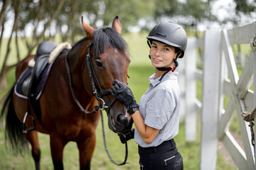 Portrait of female horseman with her brown Thoroughbred horse outdoors. Concept of animal care. Rural rest and leisure. Idea of green tourism. Green countryside landscape on sunny day