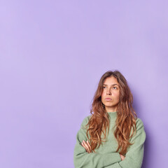 Wall Mural - Thoughtful beautiful young woman keeps arms folded concentrated above thinks about something wears casual jumper isolated over purple background with copy space for your advertising content.