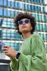 Wall Mural - Outdoor shot of curly haired woman wears sunglasses and green jumper uses smartphone while walking at street among modern glass city building. Female blogger spends free day outside uses internet