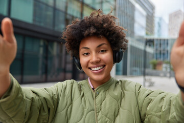 Sticker - Positive curly haired woman smiles happily makes selfie listens music via headphones wears jacket poses outdoors against urban background spends free time in big city. People lifestyle hobby