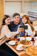 © Andrii - A group of young cheerful friends is sitting in a cafe talking and taking selfies on the phone. Lunch at the pizzeria.