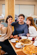 © Andrii - A group of young cheerful friends is sitting in a cafe talking and taking selfies on the phone. Lunch at the pizzeria.